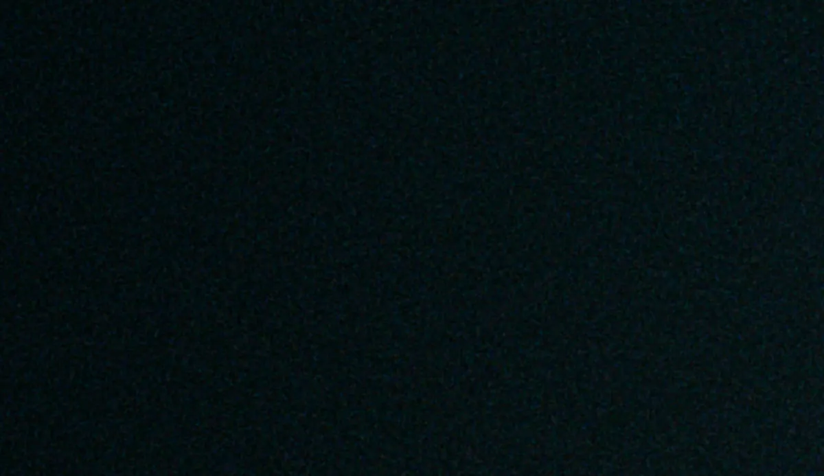 Production still — a man’s eye peering through a narrow opening of light in a dark doorway.